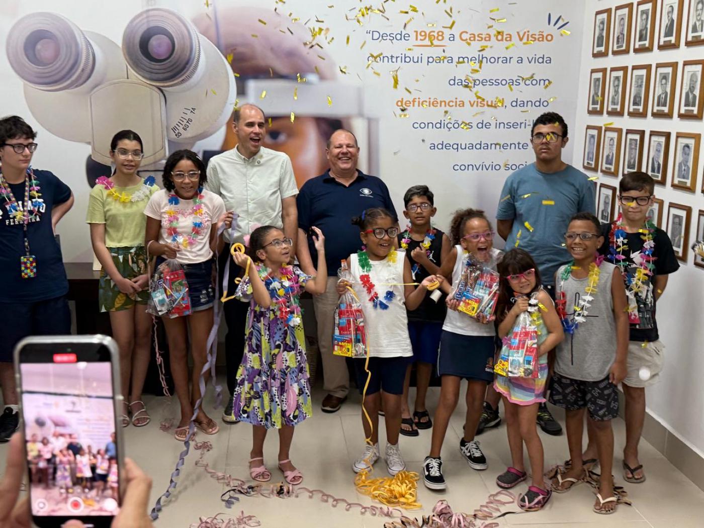crianças usando óculos posam para foto com alguns adultos. O ambiente está decorado com itens de carnaval. #paratodosverem