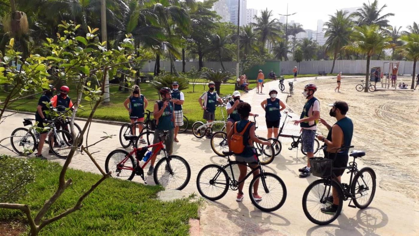 Grupo de ciclistas está reunido, parado, no jardim da orla. #Paratodosverem
