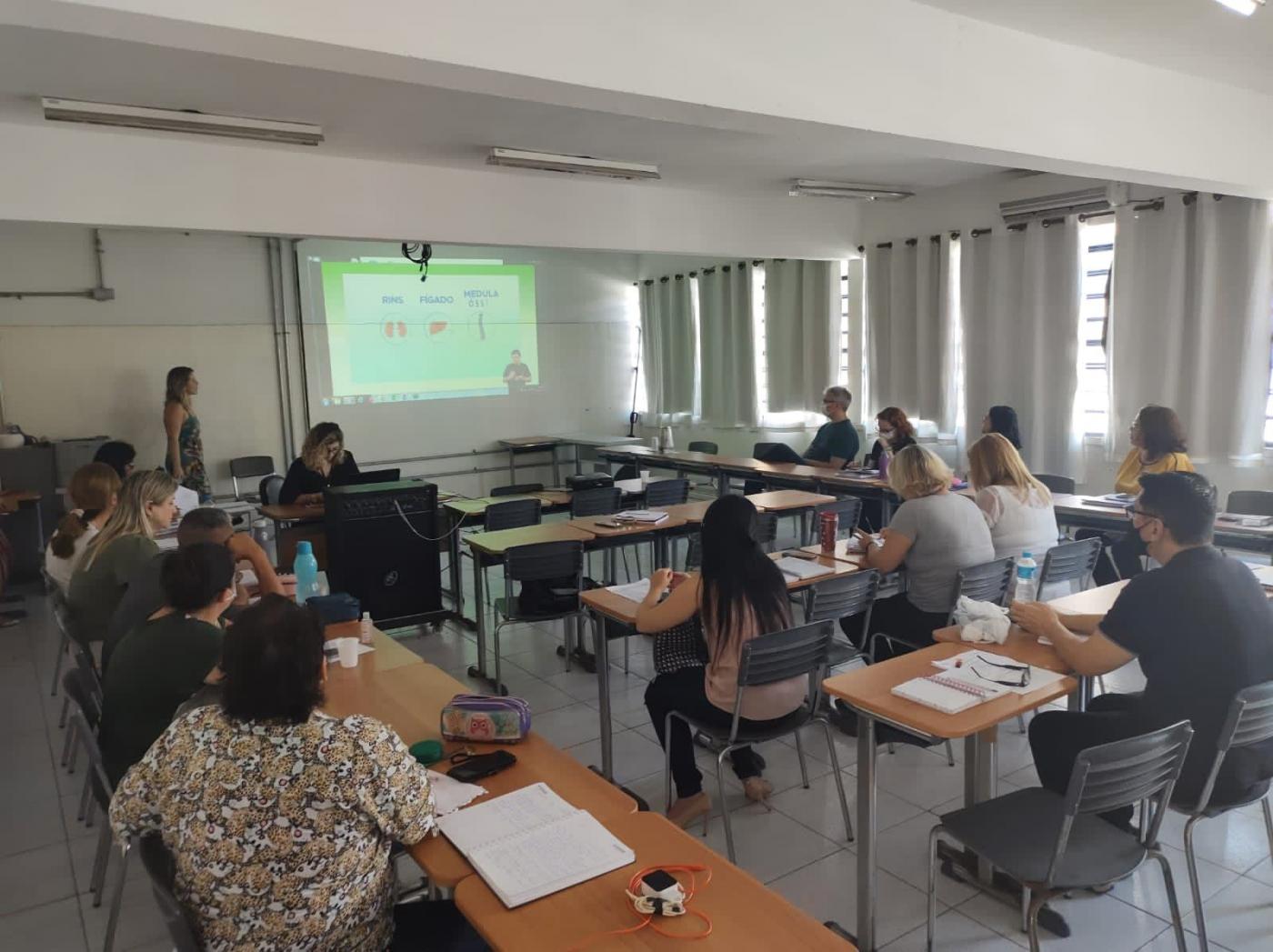 sala de aula com adultos sentados assistindo a uma mulher que fala à frente de todos e diante de uma tela onde estão projetadas imagens sobre doação de órgãos. #paratodosverem