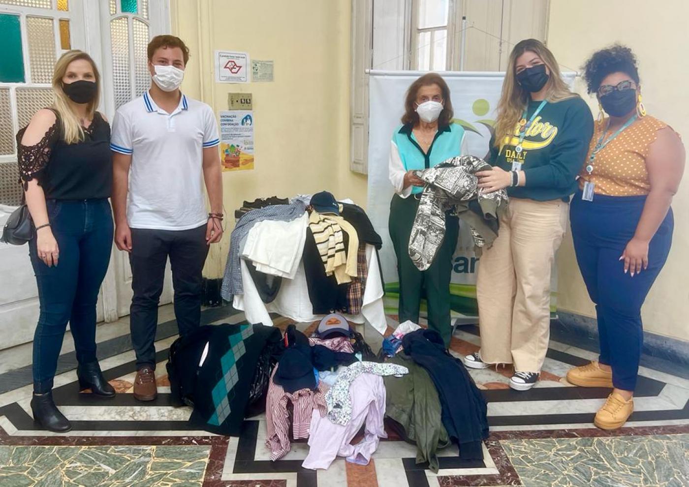 roupas dentro de caixas no chão. Cinco pessoas posam para foto atrás das caixas. #paratodosverem