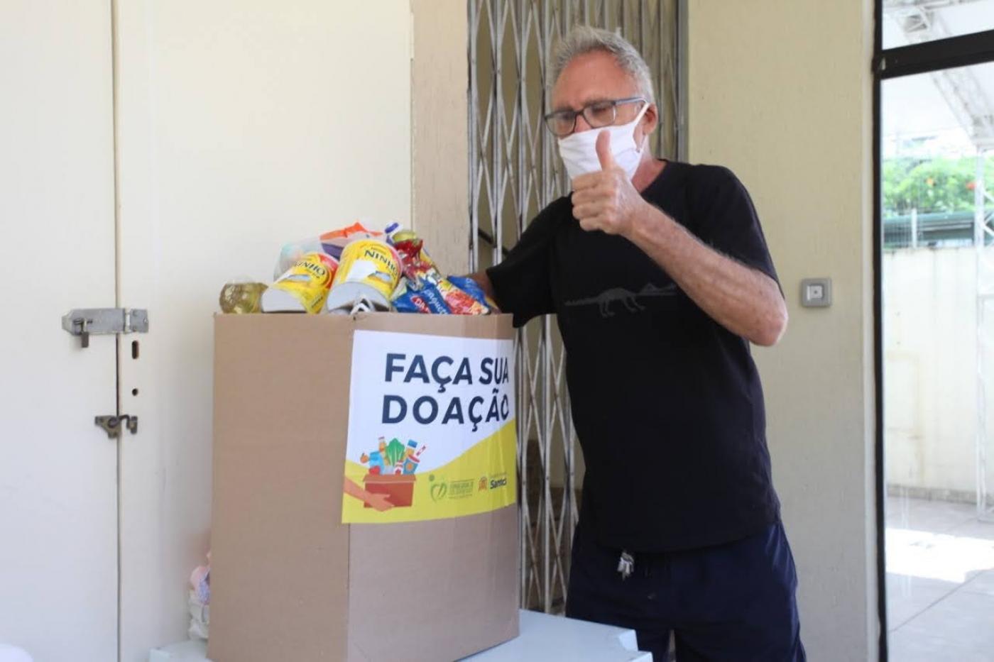 Homem usando máscara deposita alimento em caixa de papelão. Na frente da caixa está escrito 