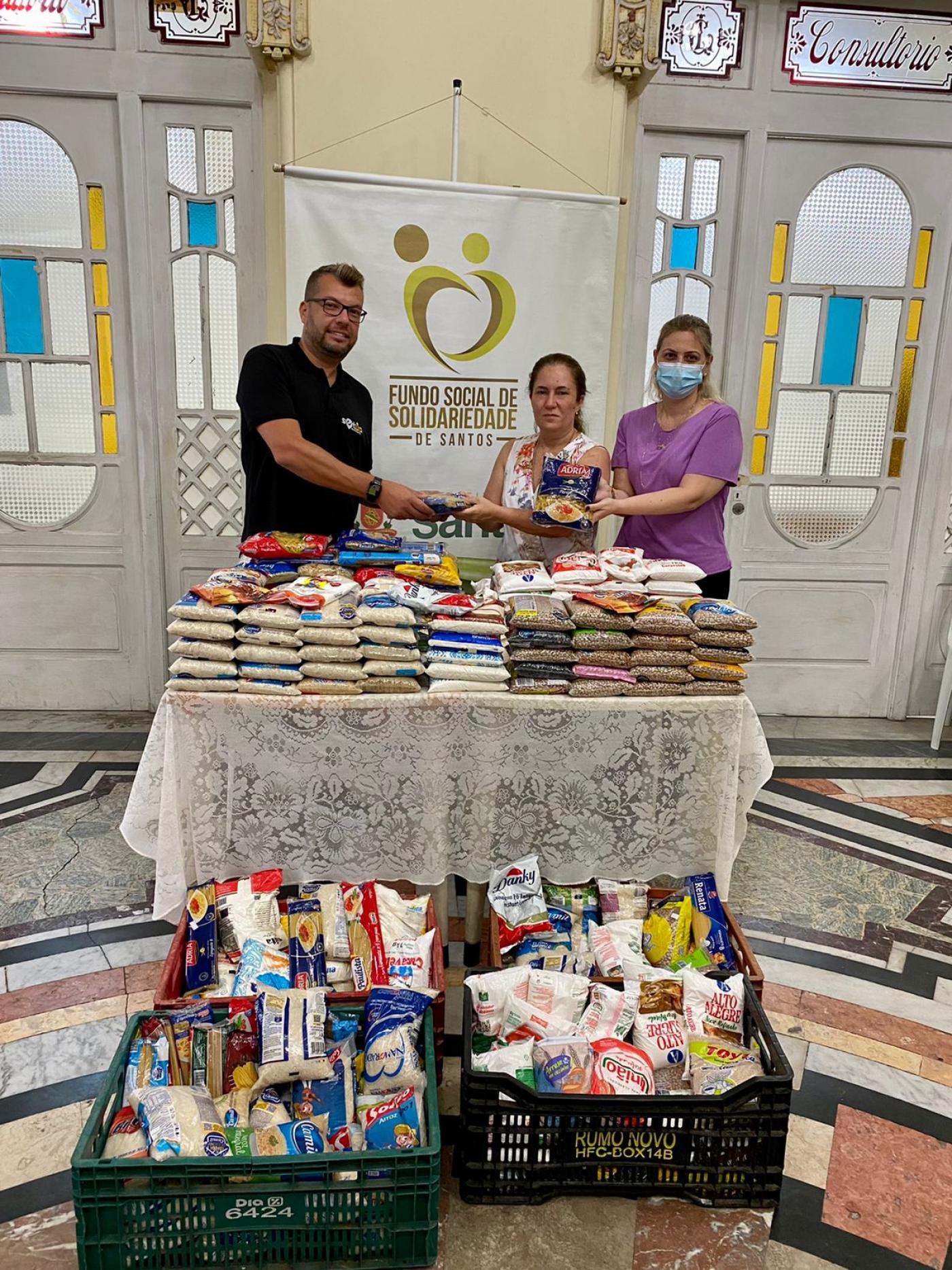 um homem e duas mulheres estão atrás de uma mesa cheia de pacotes de alimentos. No chão também há duas caixas cheias de alimentos. Ao fundo, um banner do fundo social de solidariedade. #paratodosverem