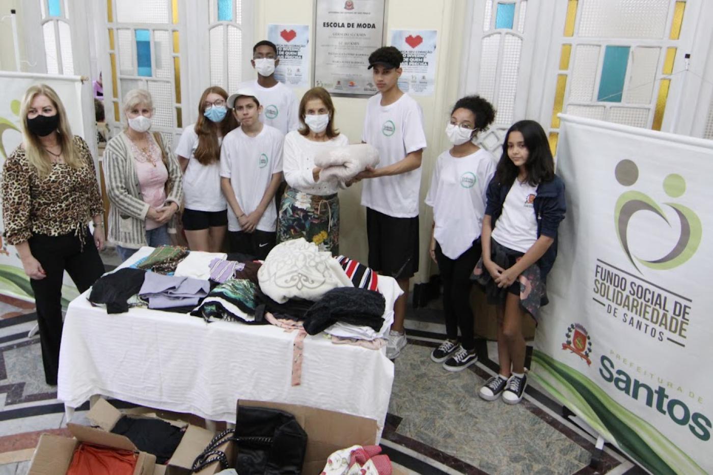 mesa cheia de roupas e cobertores com estudantes e adultos atrás posando para a foto. Há um banner do fundo social de solidariedade 