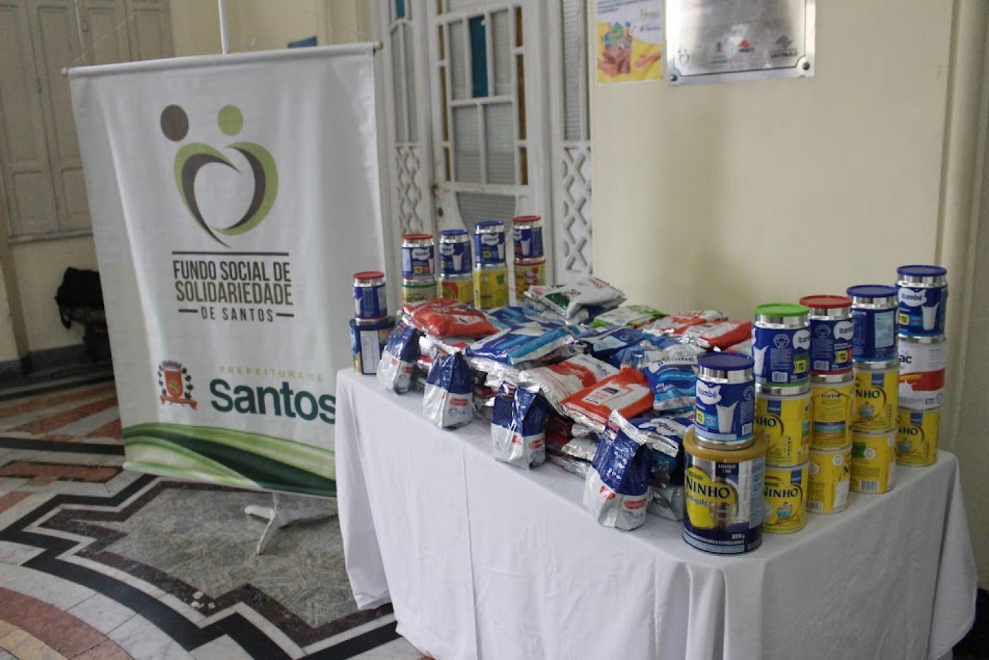 pacotes e latas de leite em pó sobre uma mesa. Ao lado, um banner do fundo social de solidariedade. #paratodosverem