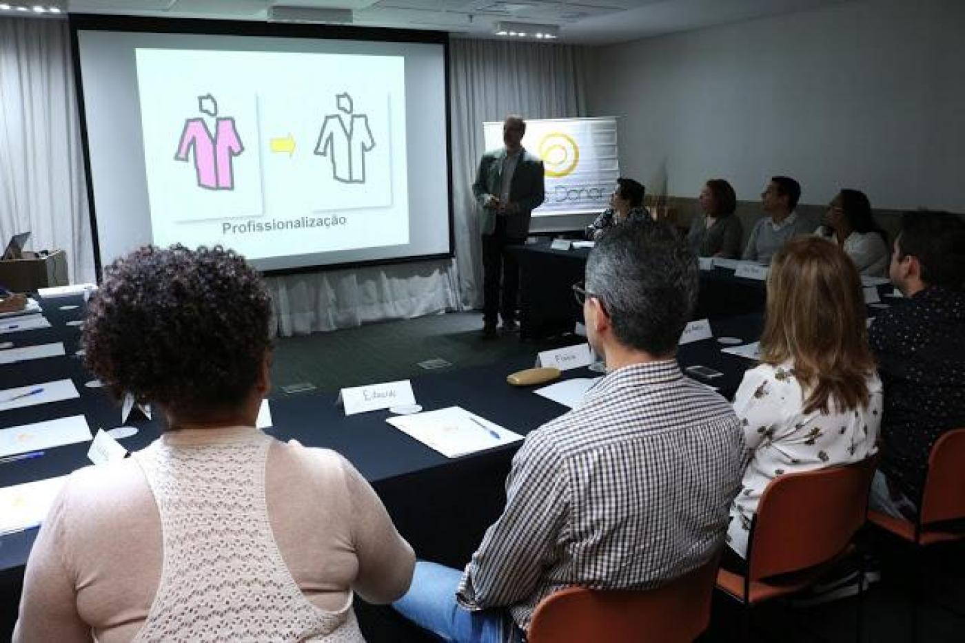 Profissionais estão reunidos em sala numa mesa em forma de U. Ao fundo um homem fala ao grupo. Há um telão que projeto a imagem de dois corpos desenhados. #Pracegover