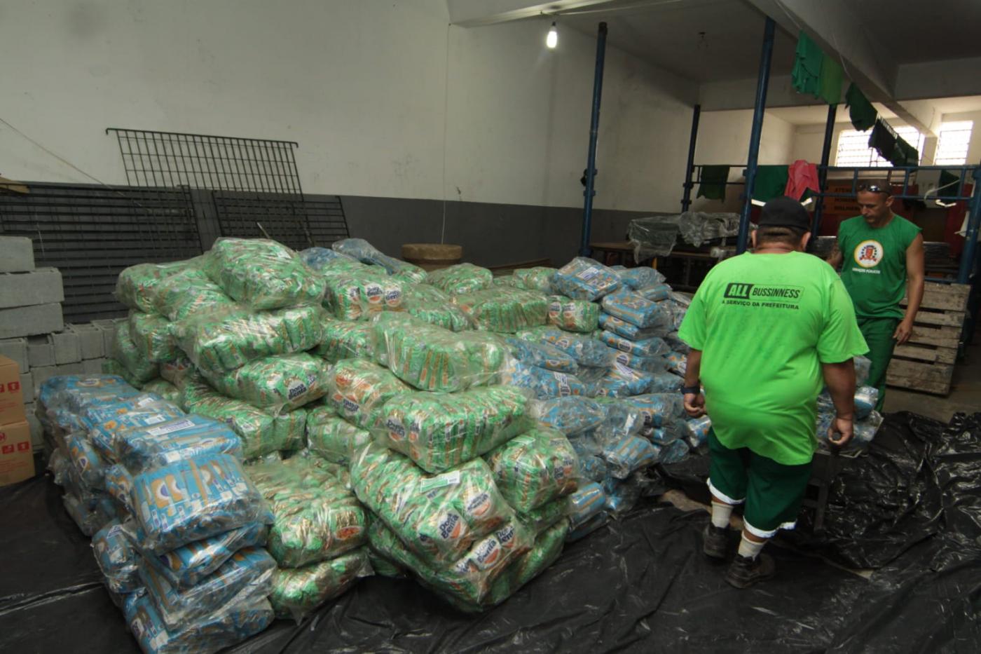 Alimentos empilhados em galpão com dois trabalhadores ao lado. #pratodosverem