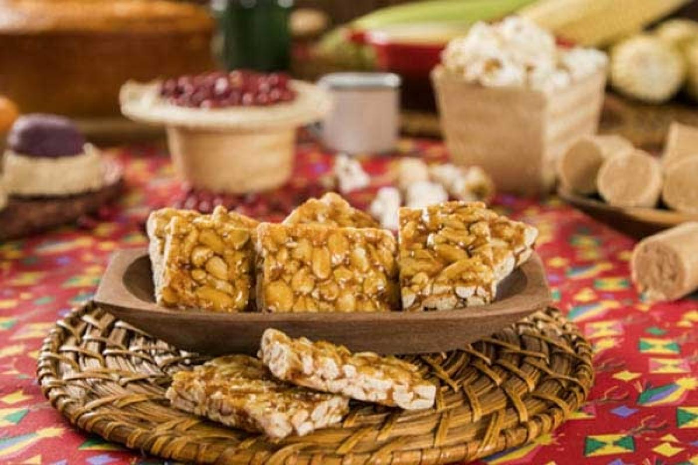 Mesa com doces como pé de moleque e paçoca. Amendoim e pipoca ao fundo. #paratodosverem