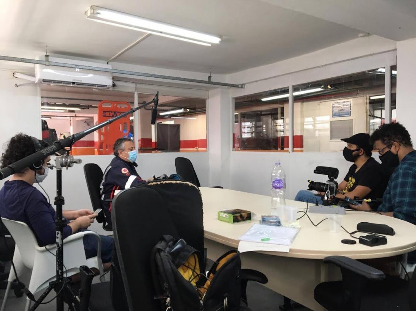 Sala com equipe de gravação. Há um microfone montado, dois homens fazendo a filmagem e um sendo entrevistado. #Paratodosverem