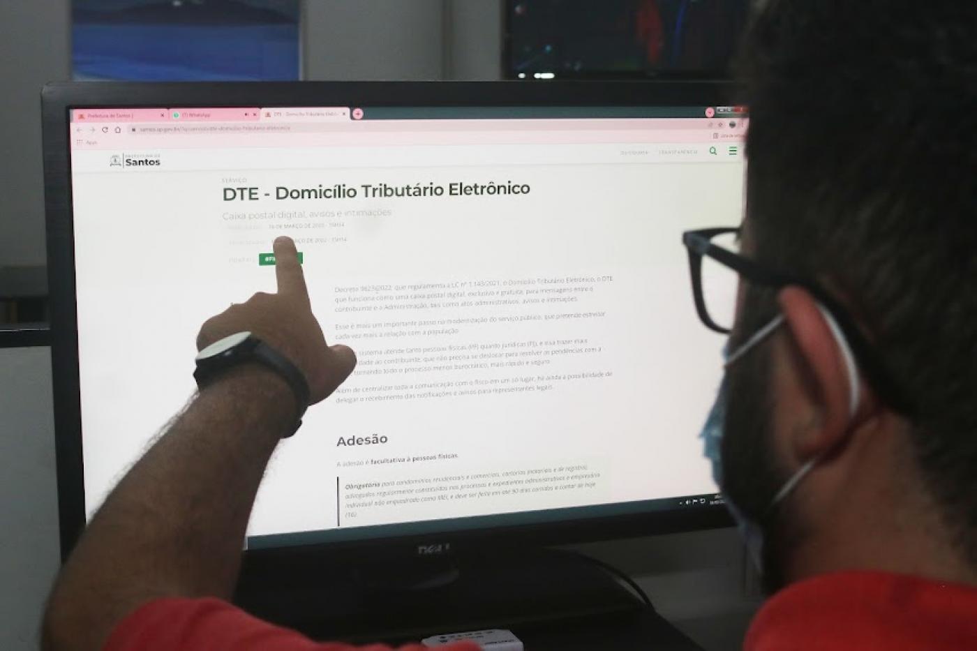 homem está de costas para foto apontando dedo para tela de desktop aberto n a página do Domicílio eletrônico. #paratodosverem