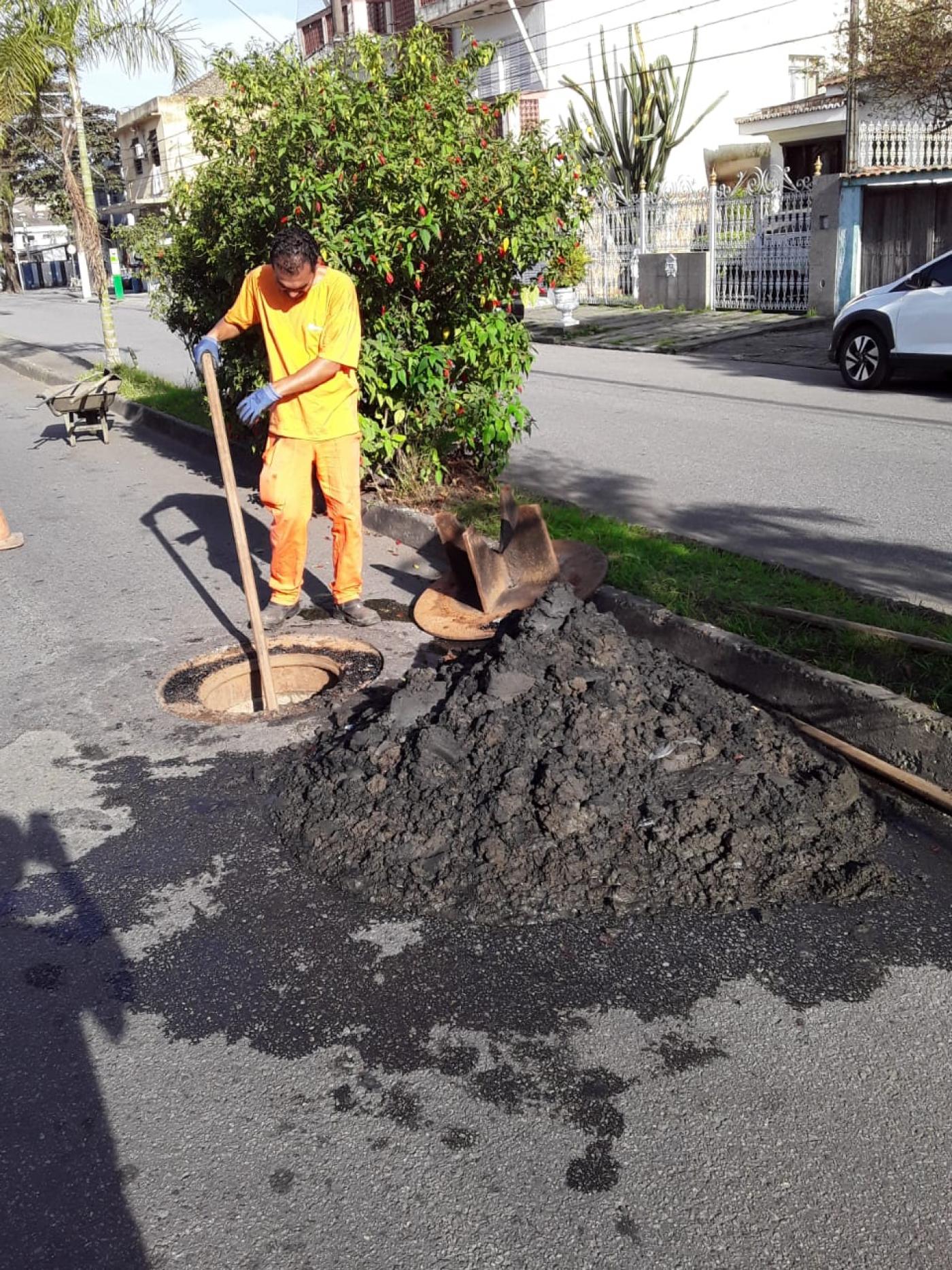 operário uniformizado segura uma longa ferramenta que está dentro de um poço de visita em Santos. Ao lado, na pista há um acumulado de lama. #paratodosverem