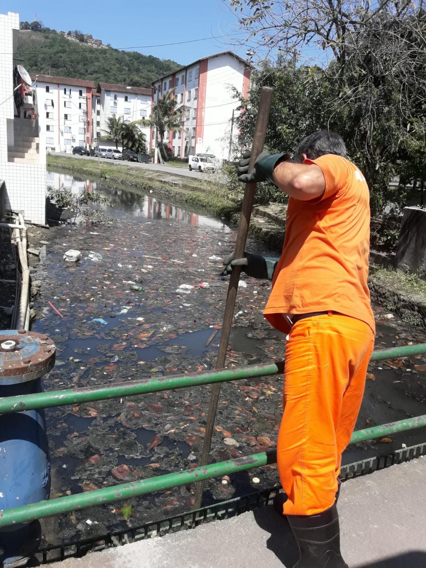 homem limpa trecho de canal, do lado de fora, com auxílio de uma ferramenta de cabo longo