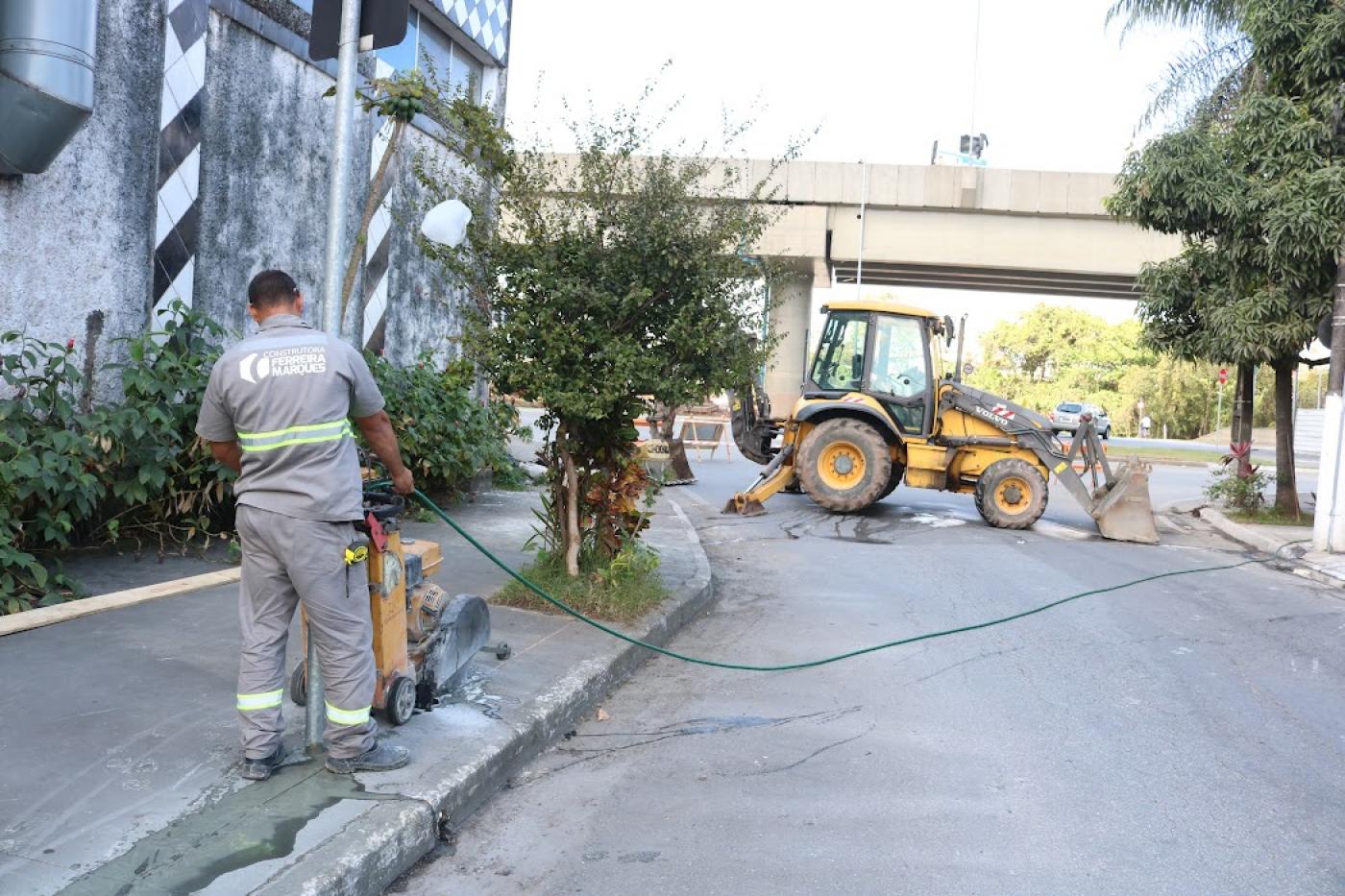 Profissional trabalha em obra de sarjeta com retroescavadeira ao fundo. #pratodosverem