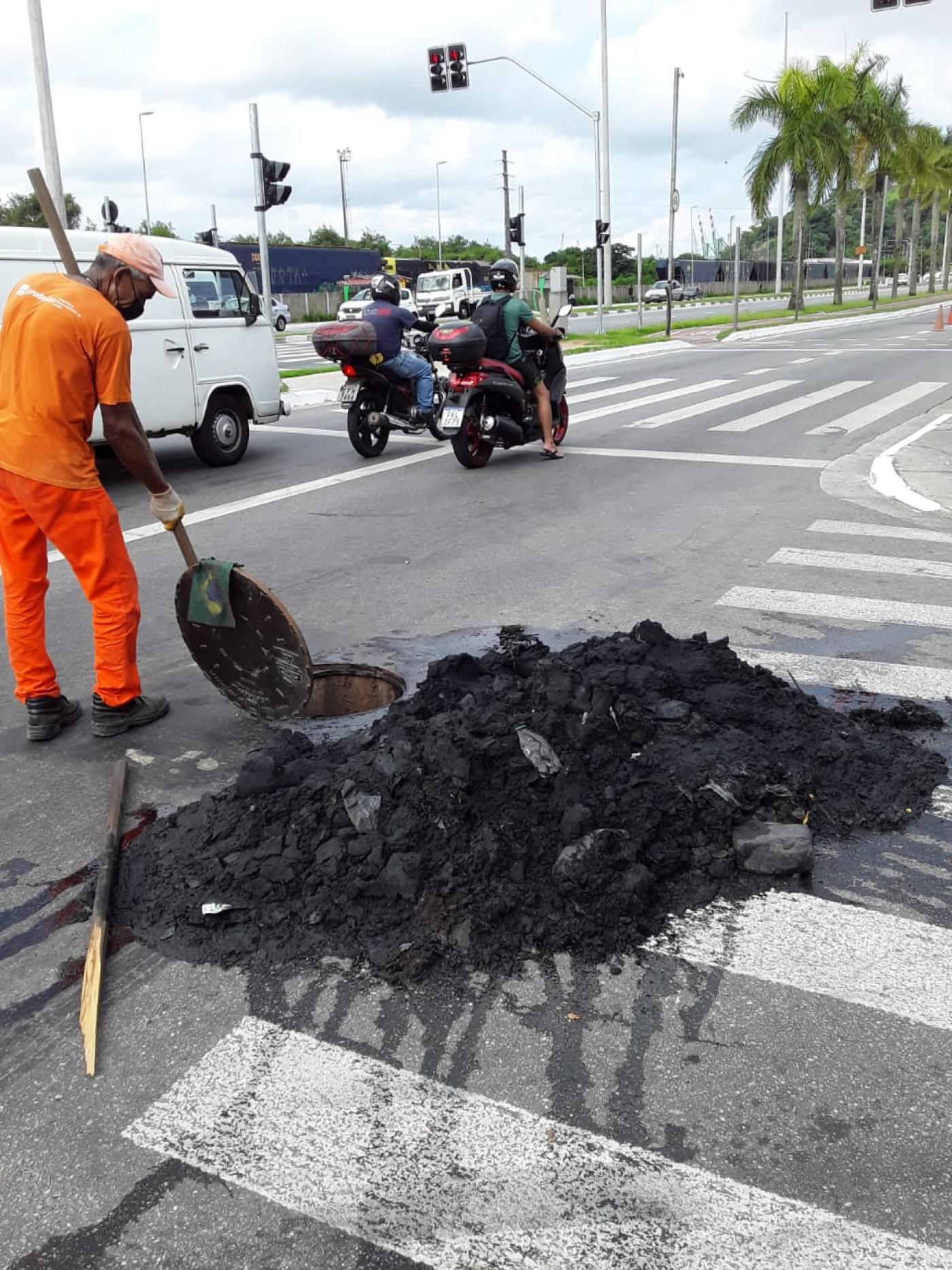 Homem está segurando pá ao lado de um monte de resíduos retirados do sistema de drenagem. #Paratodosverem