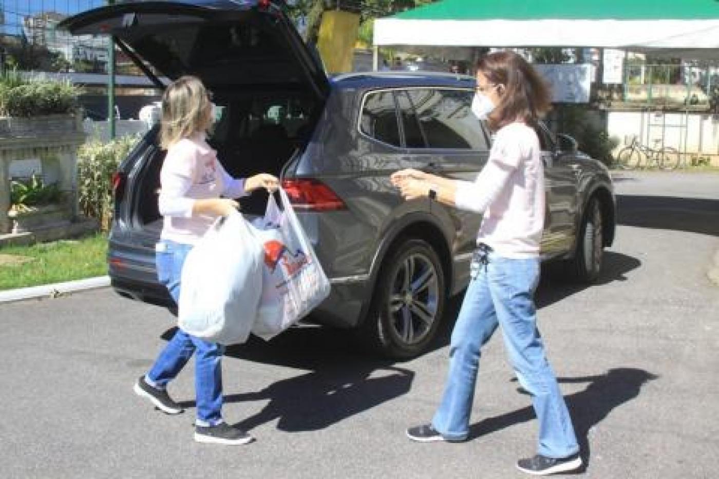 Mulher entrega sacolas com doações para voluntária, com carro ao fundo. 