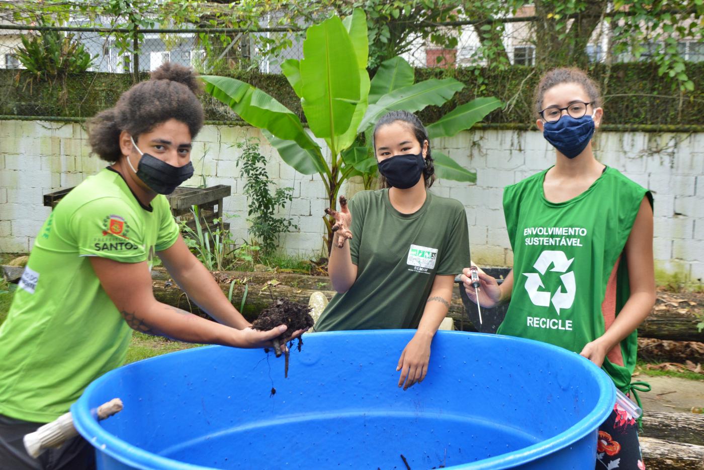 Três jovens seguram terra e local de compostagem #paratodosverem