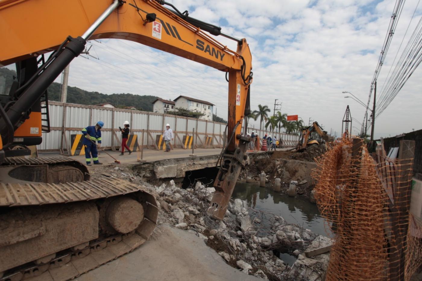 Máquinas e homens trabalham na construção de pontilhão. A máquina está removendo parte do piso. Ao redor se vê um trecho de rio. #Pracegover