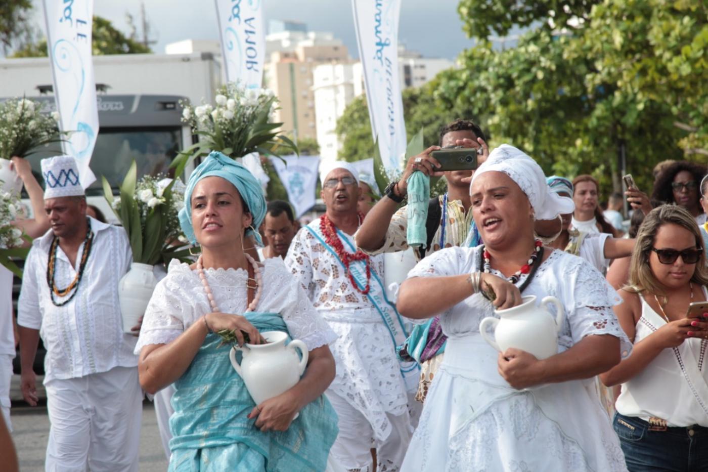 Mulheres vestidas com roupas típicas de umbanda, com turbantes e vasos nas mãos caminham pela praia. #Pracegover