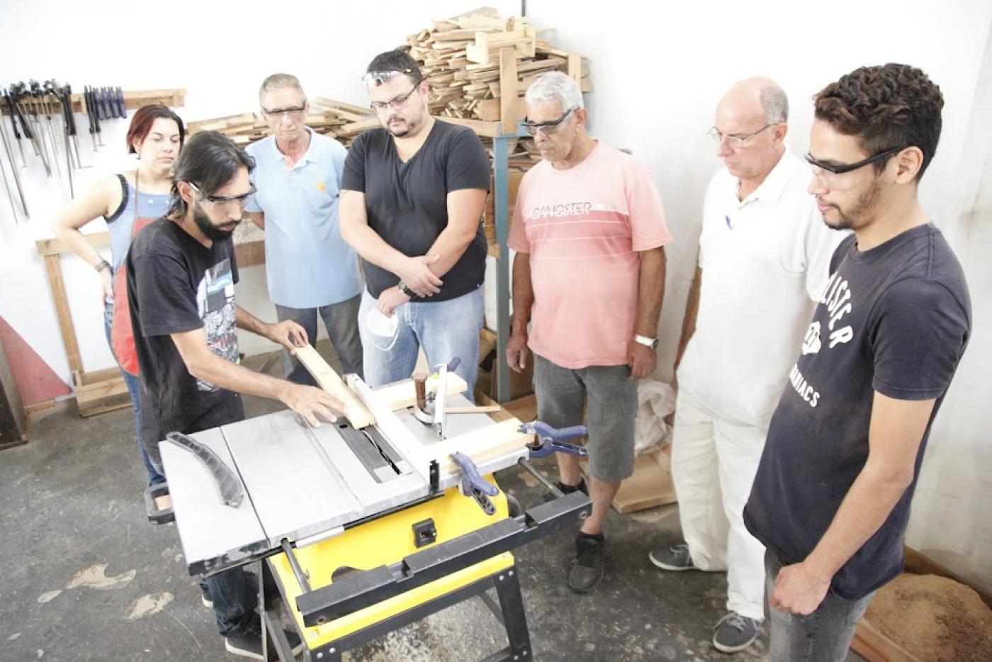 homem está manejando máquina de corte com pedaço de madeira em cima. Ele é observado por seis pessoas em torno dele. Ambiente tem madeiras empilhadas ao fundo. #paratodosverem