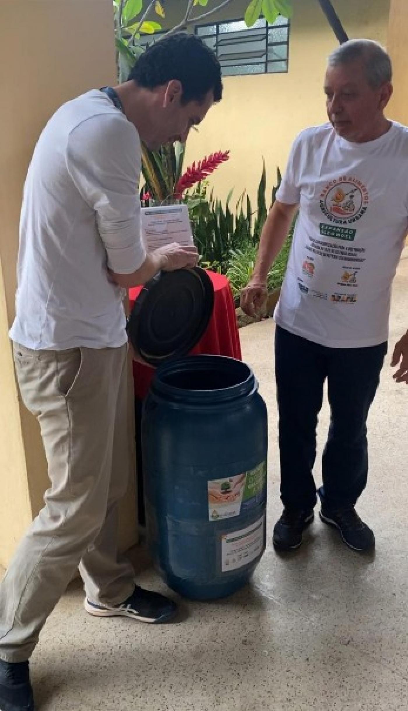 dois homens em pé ladeiam tonel para depósito de óleo de cozinha usado. #paratodosverem