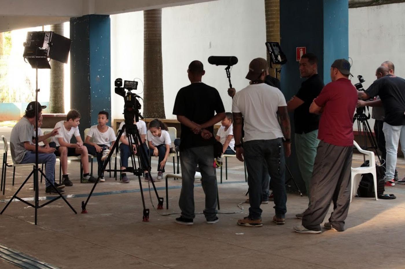 equipe de produção visual, com câmeras está dinate de de crianças que estão sentadas em cadeiras dispostas lado a lado num grande salão. #paratodosverem
