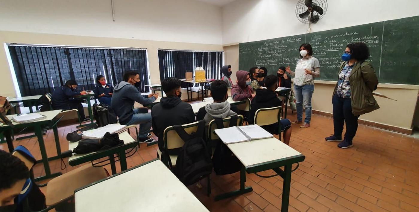 sala de aula com alunos sentados e duas mulheres em pé. Uma delas fala ao grupo. #paratodosverem