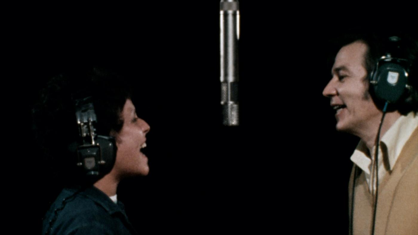Eis e tom gravam música ao microfone. Eles estão frente a frente. #paratodosverem