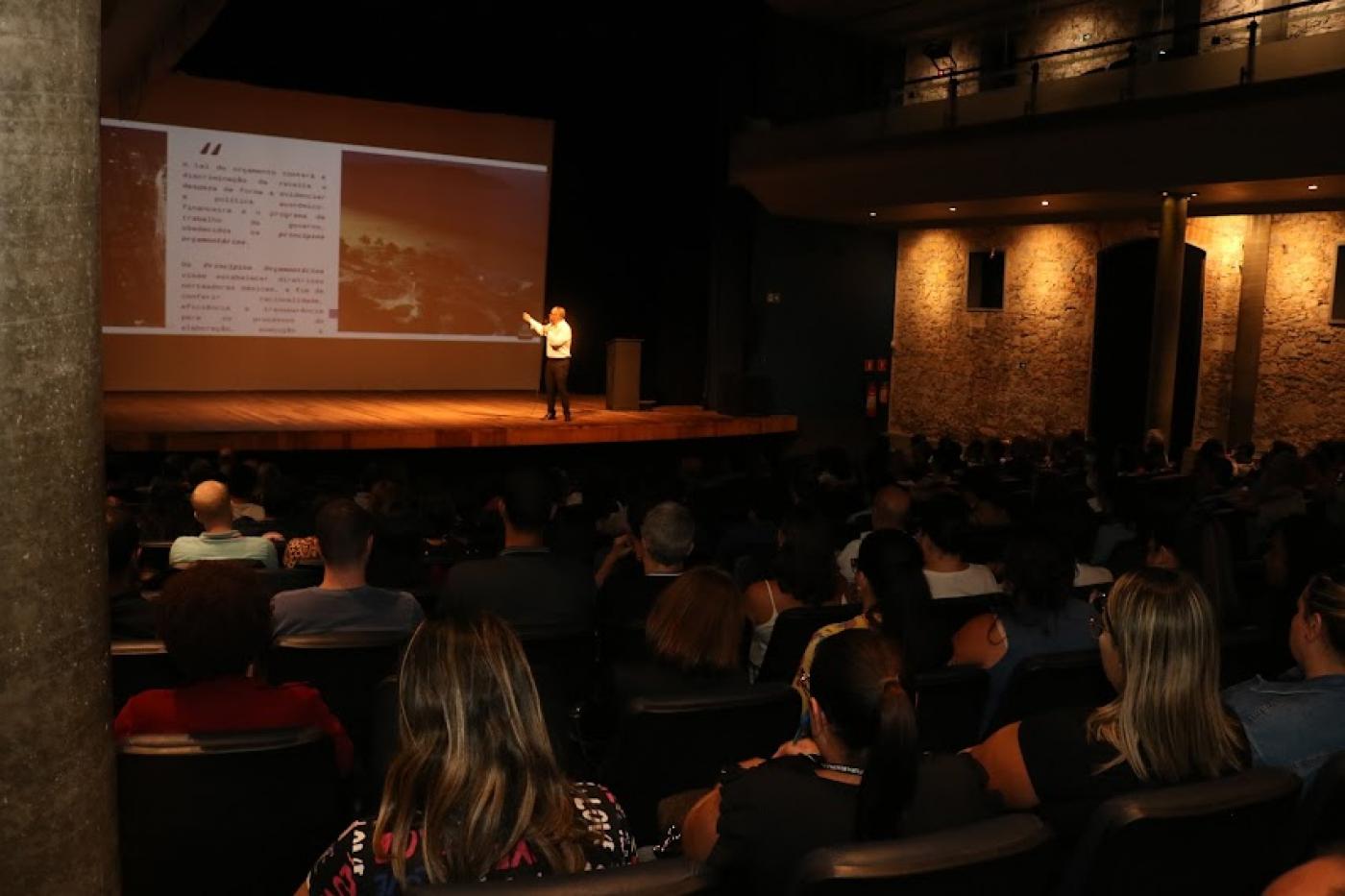 Servidores sentados na plateia do teatro assistem exibição de secretário, em pé no palco, abaixo de telão. #pratodosverem