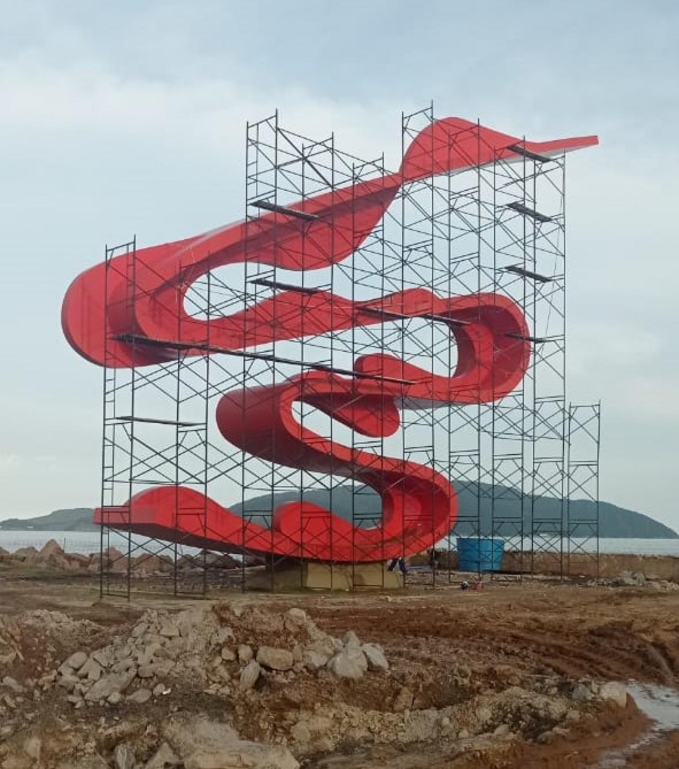 escultura cercada por andaimes, com areia na frente e mar ao fundo #paratodosverem