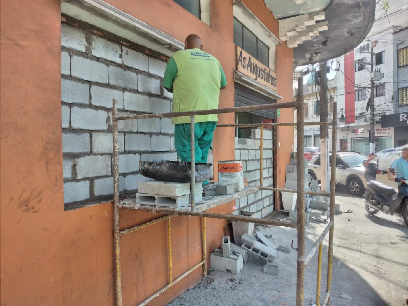 operário fecha janela de imóvel com blocos. Ele está sobre um andaime. #paratodosverem