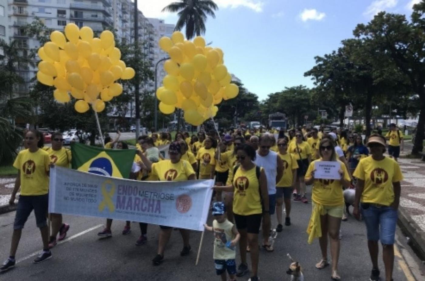 pessoas caminhando na avenida, usando roupas amarelas. Algumas seguram balões amarelos em alusão à endometriose. Duas pessoas à frente da marcha seguram uma faixa onde se lê Endomarcha. #paratodosverem