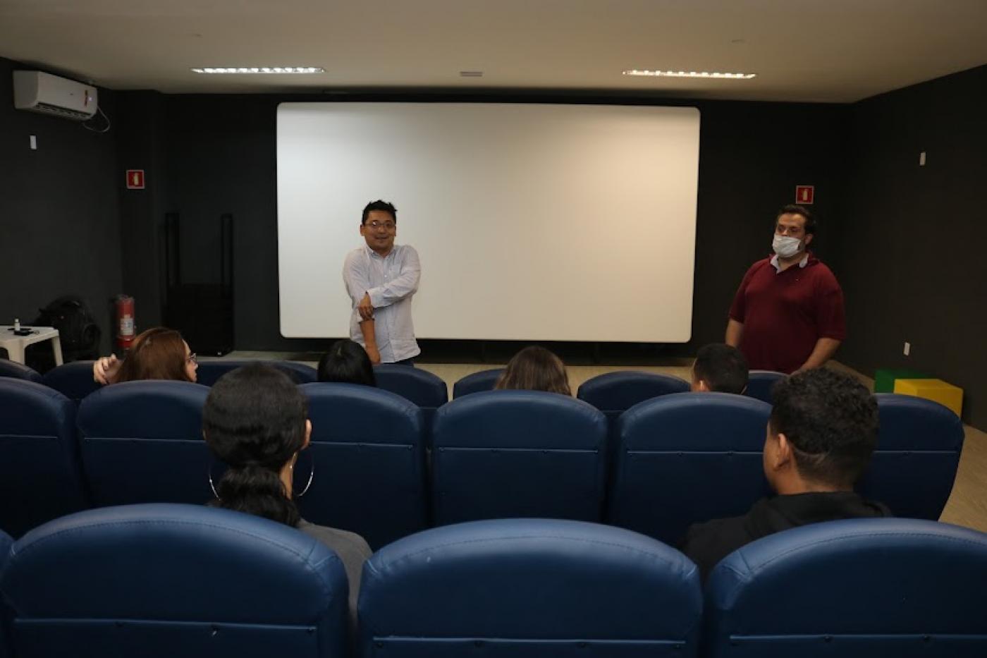 jovens estão sentados em poltrona de auditório atentes a dois homens que estão em pé falando. Ao fundo, um telão em branco. #paratodosverem