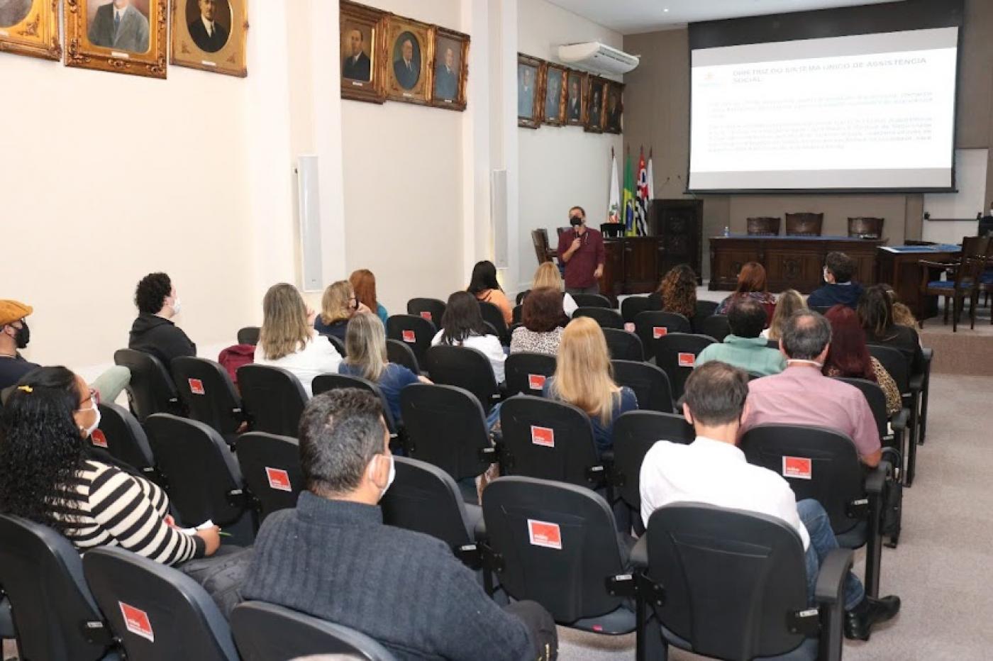 Pessoas sentadas em um auditório observam pessoa falando e telão ao fundo #paratodosverem