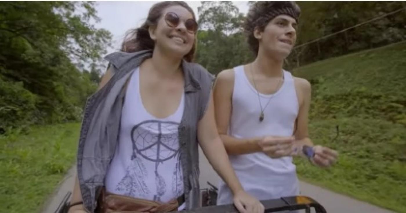 cena do filme. Mulher e um rapaz estão em uma estrada. Eles estão em pé dentro de um carro aberto. Não se vê o carro, somente parte da estrutura superior onde a mulher pousa as mãos. #paratodosverem 