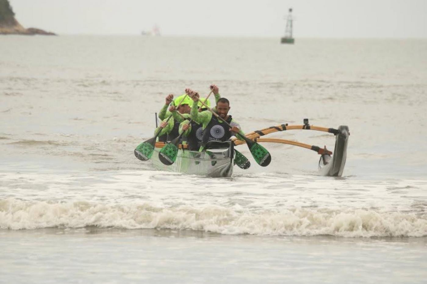 equipe em canoa havaiana vem chegando na beira da praia. Ao fundo, apenas o farol no meio do mar. #paratodosverem