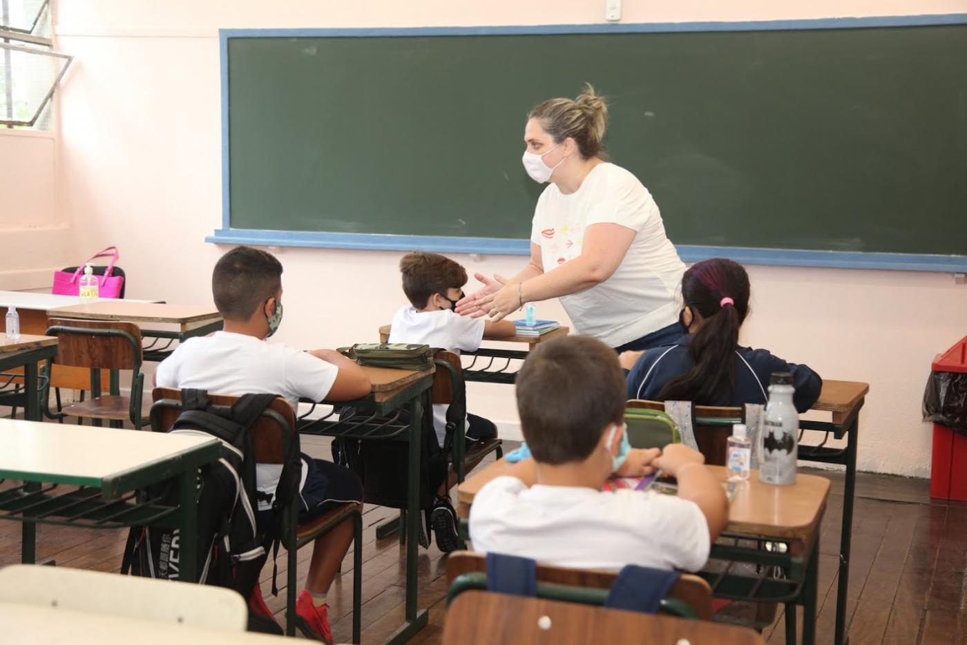 Sala de aula com professora à frente dos alunos sentados em suas carteiras. #Paratodosverem