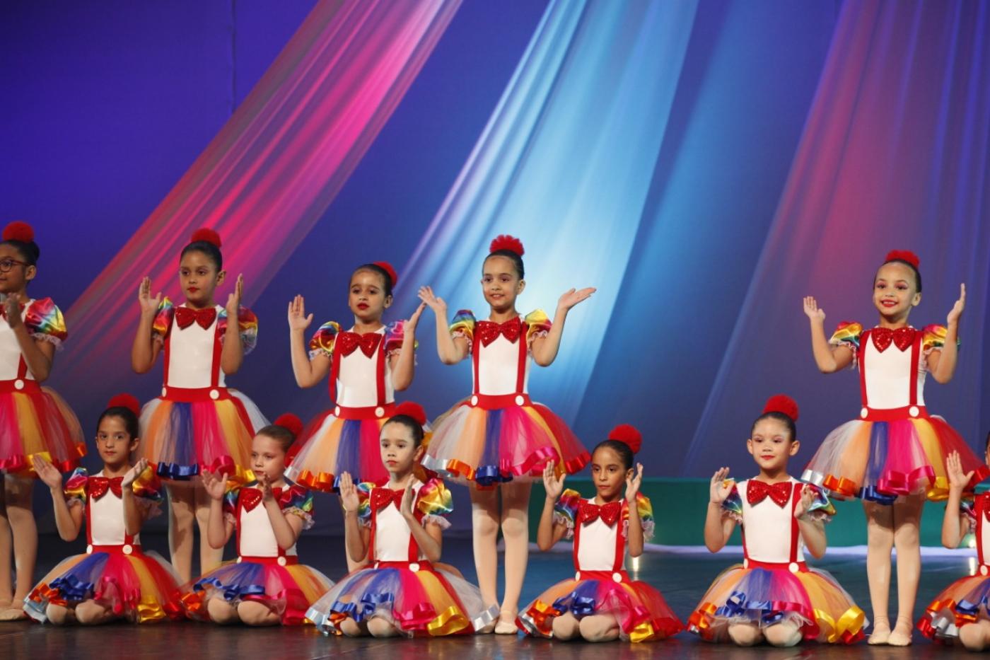 meninas estão usando a mesma roupa em apresentação de balé. Algumas estão sentadas e outras em pé no palco. #paratodosverem