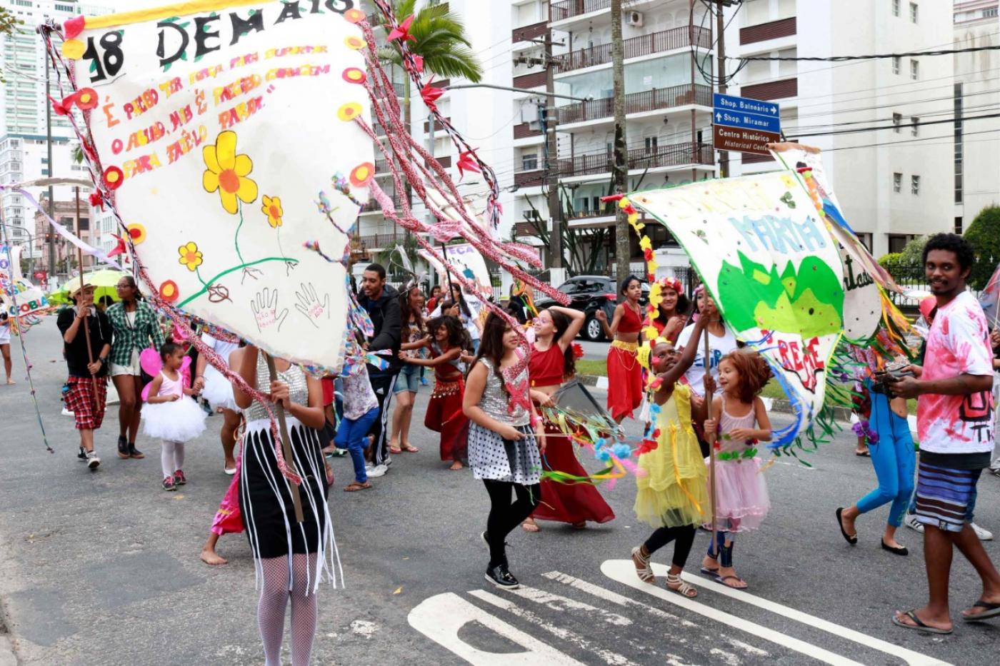 Crianças desfilam em avenida. Elas carregam estandartes e vestem roupas coloridas. #Pracegover