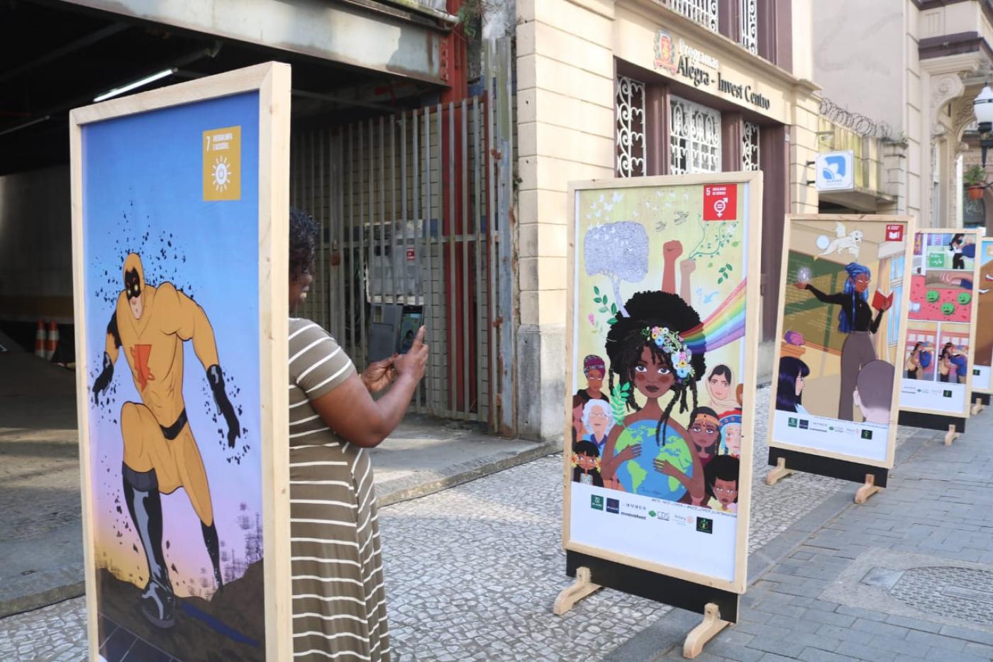 paineis expostos em calçadão. Atrás de um deles aparece a lateral do corpo de uma mulher que fotografa uma das obras. #paratodosverem 
