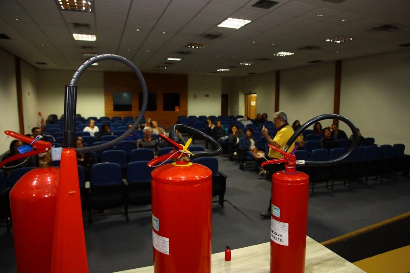 três extintores sobre uma mesa em primeiro plano. Ao fundo, um homem em pé fala a pessoas que estão sentadas em um auditório. #paratodosverem