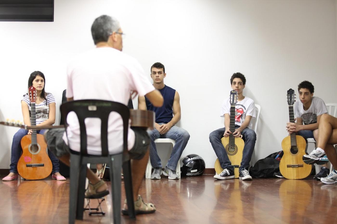 Professor sentado fala para jovens também sentados com violão na mão #paratodosverem