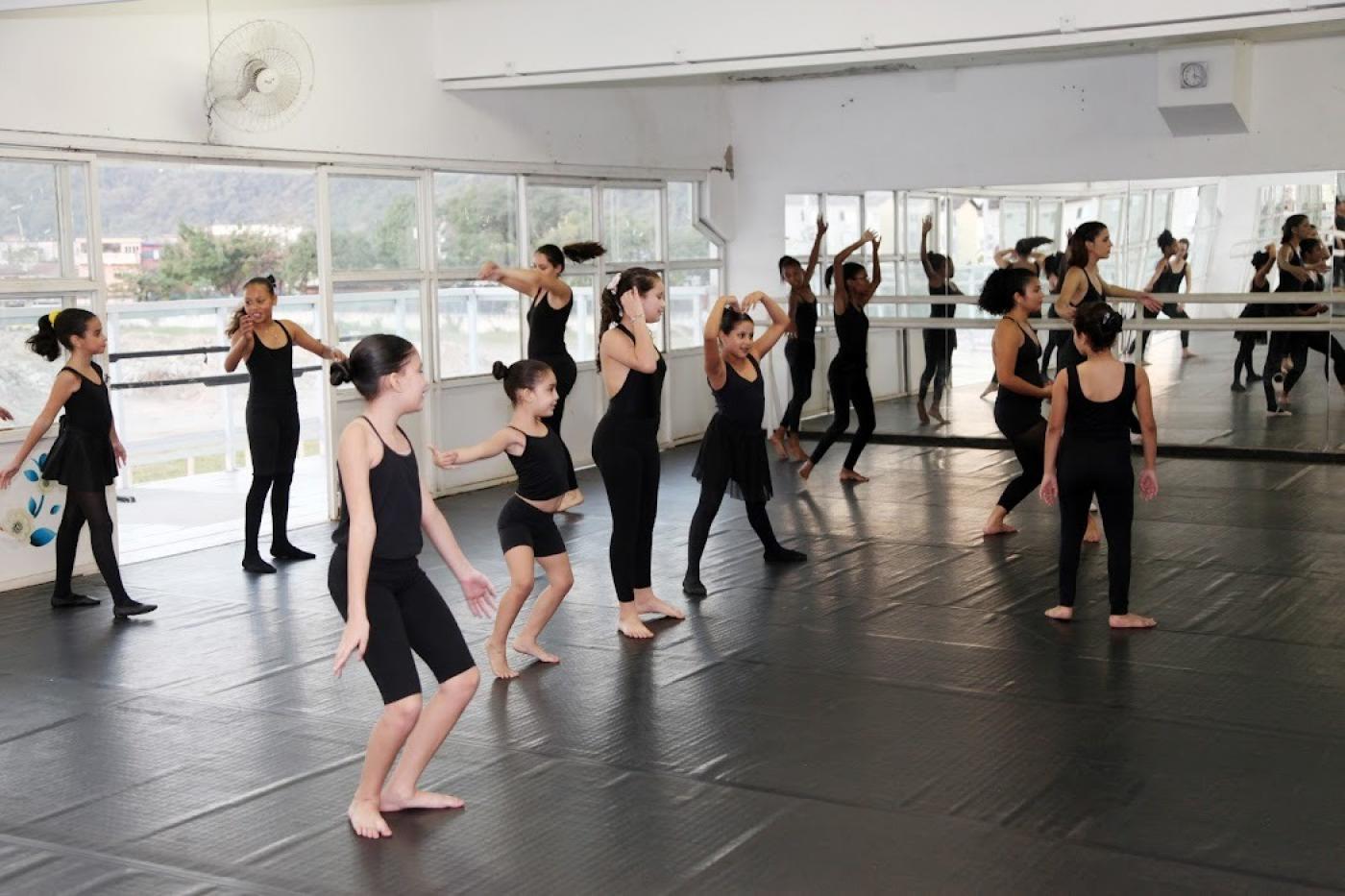 meninas estão em salão em aula de balé. Elas se movimentam em pé em diversas formas. #paratodosverem