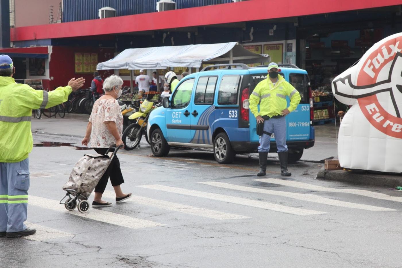 mulher atravessa rua na faixa. Ela puxa um carrinho de feira. Um agente da CET está de um lado da rua fazendo o gesto com o braço esticado. Outro está parado dooutro lado da rua. #paratodosverem