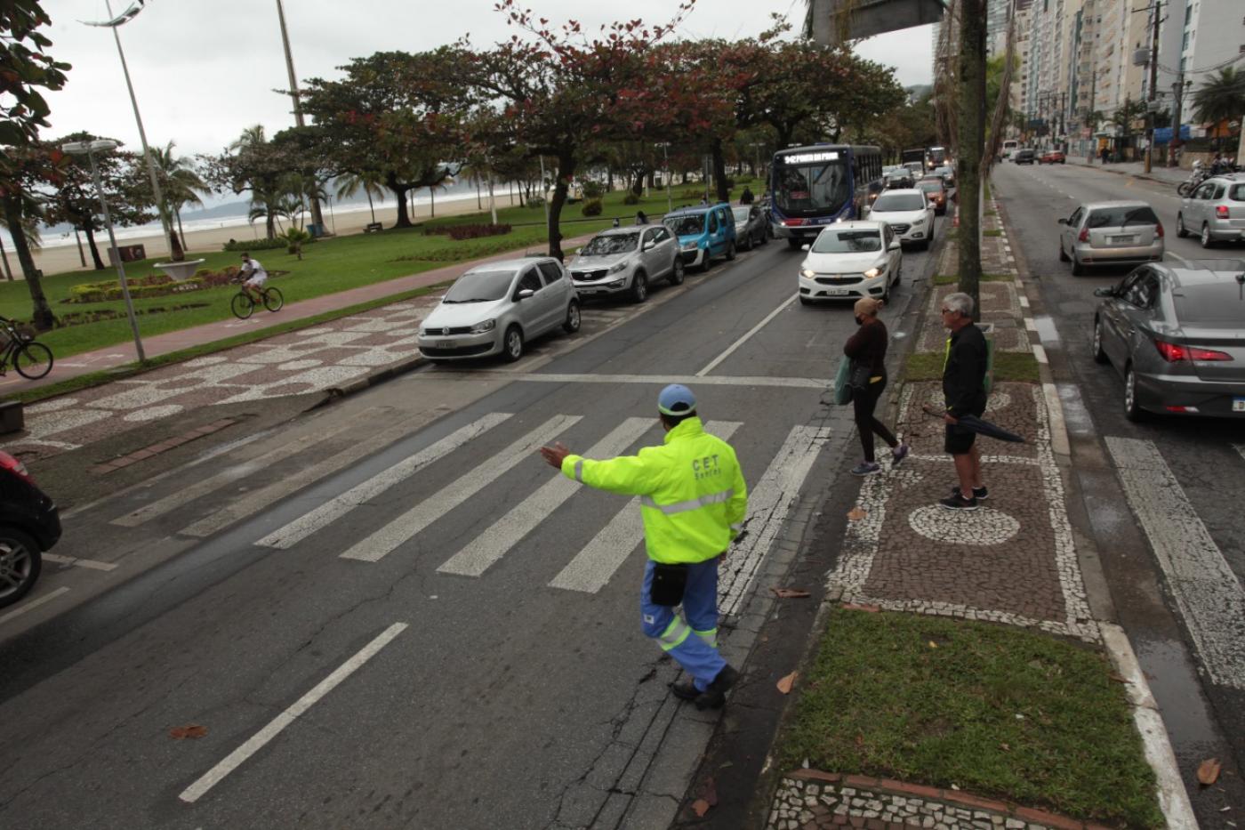 agente da CET uniformizado faz gesto com o braço para carros pararem. Duas pessoas se preparam para atravessar a avenida partindo do canteiro central. #paratodosverem
