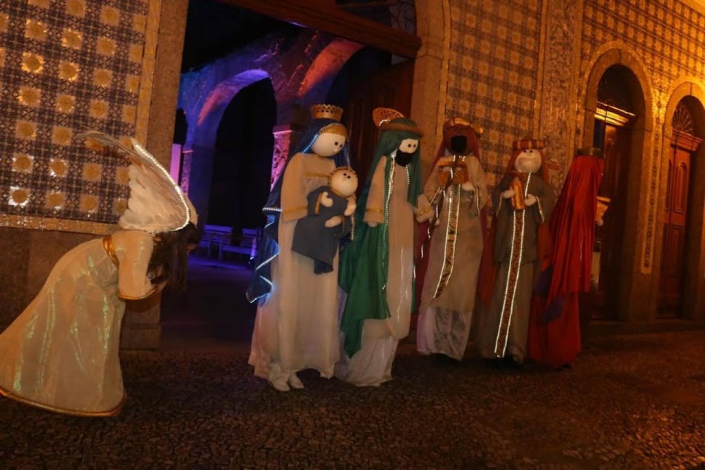 Atores vestidos de bonecos encenam Maria com o menino Jesus no colo, José, um anjo, e dois reis magos ao fundo. #paratodosverem