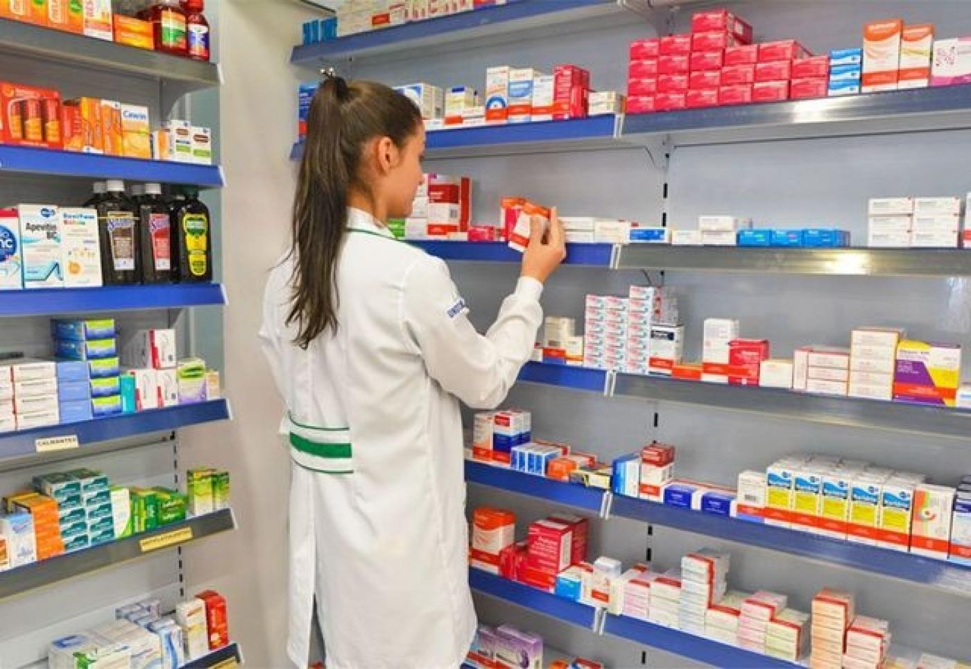 mulher usando avental está de costas para a foto e de frente  para uma prateleira cheia de remédios em uma farmácia. #paratodosverem