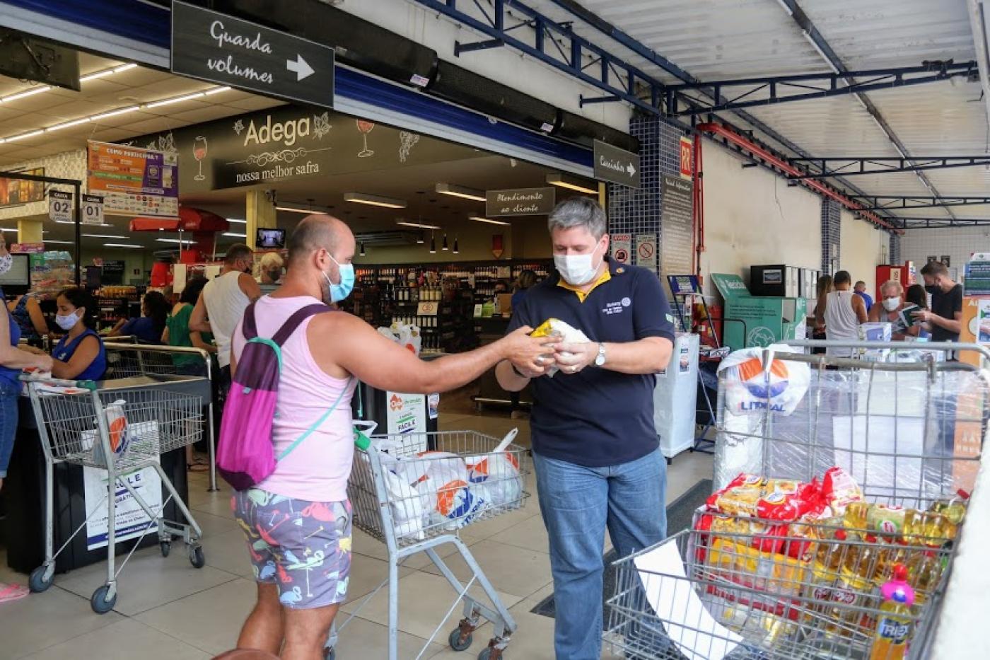 Homem de camisa branca, com carrinho de supermercado, entrega doação para homem de camisa azul em frente a supermercado. #pracegover