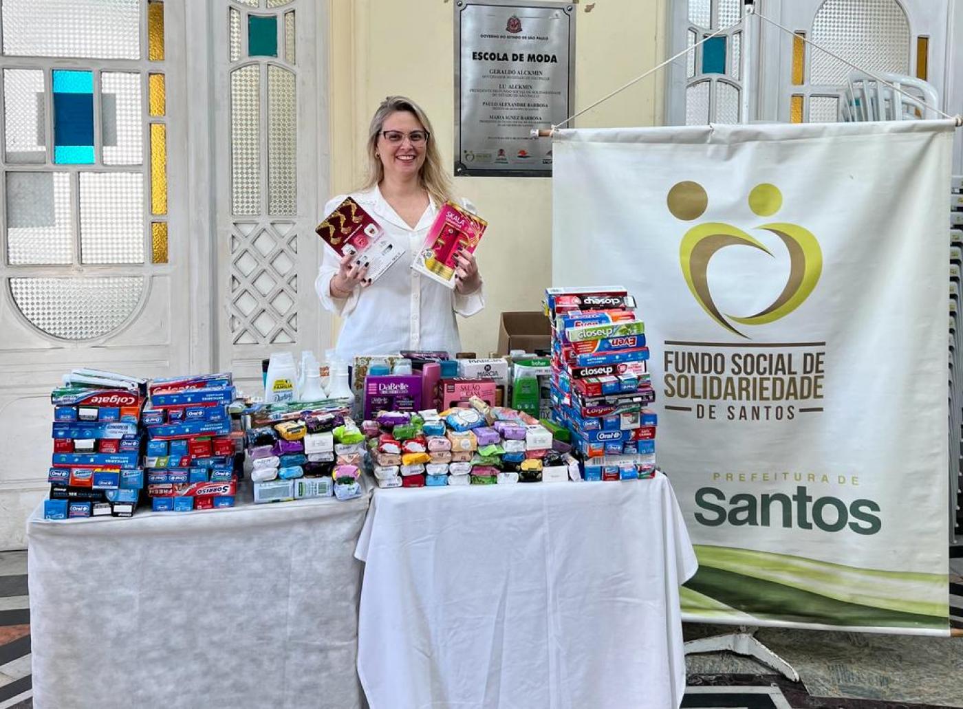 mulher segura pacote de produto de higiene e posa para foto. Diante dela há uma mesa cheia de produtos de higiene. #paratodosverem 