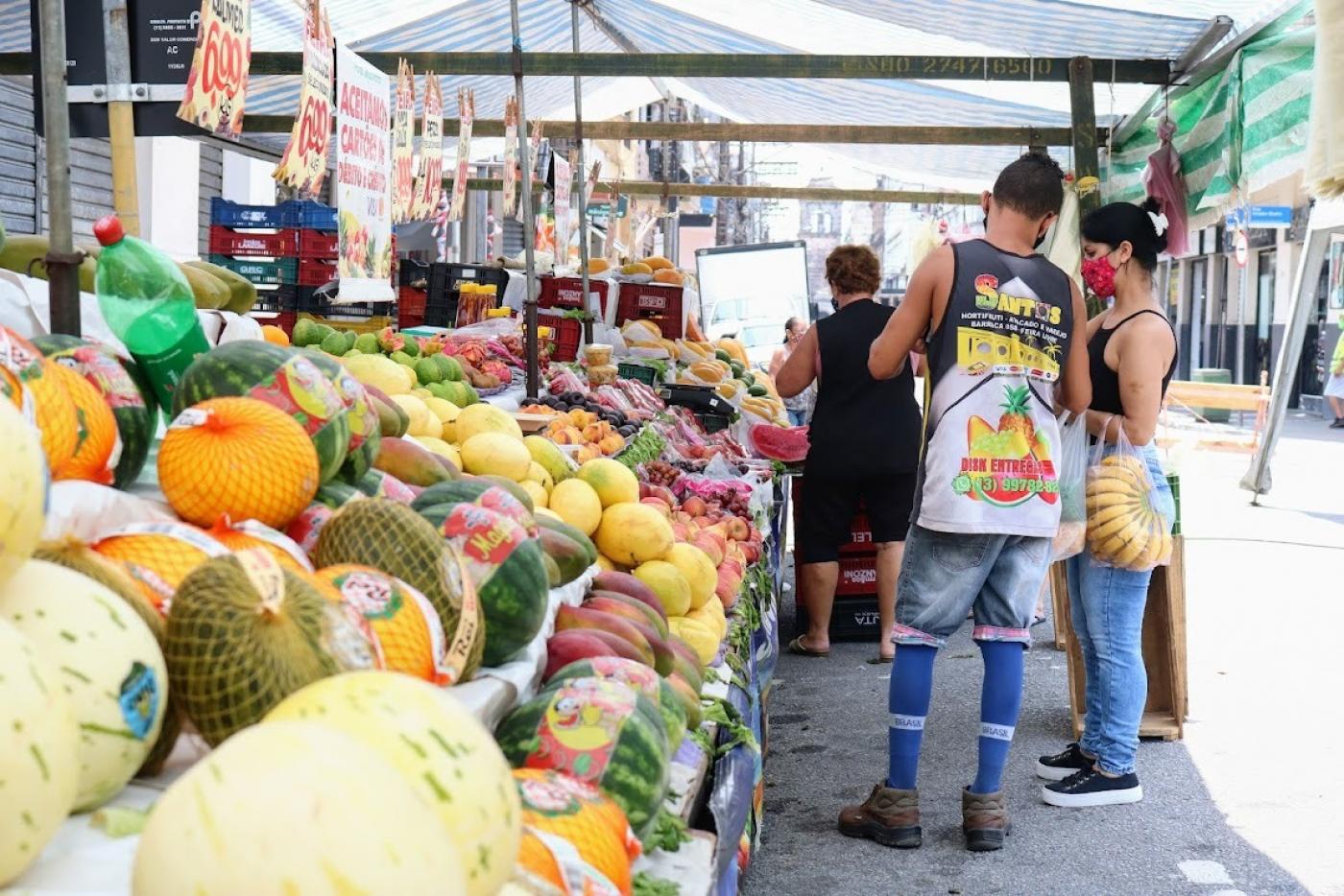 barraca de feira com frutas ao lado esquerdo. Feirante e consumidora do lado direito. Ele está de costas e ela, de lado, segurando uma sacola com bananas. #paratodosverem