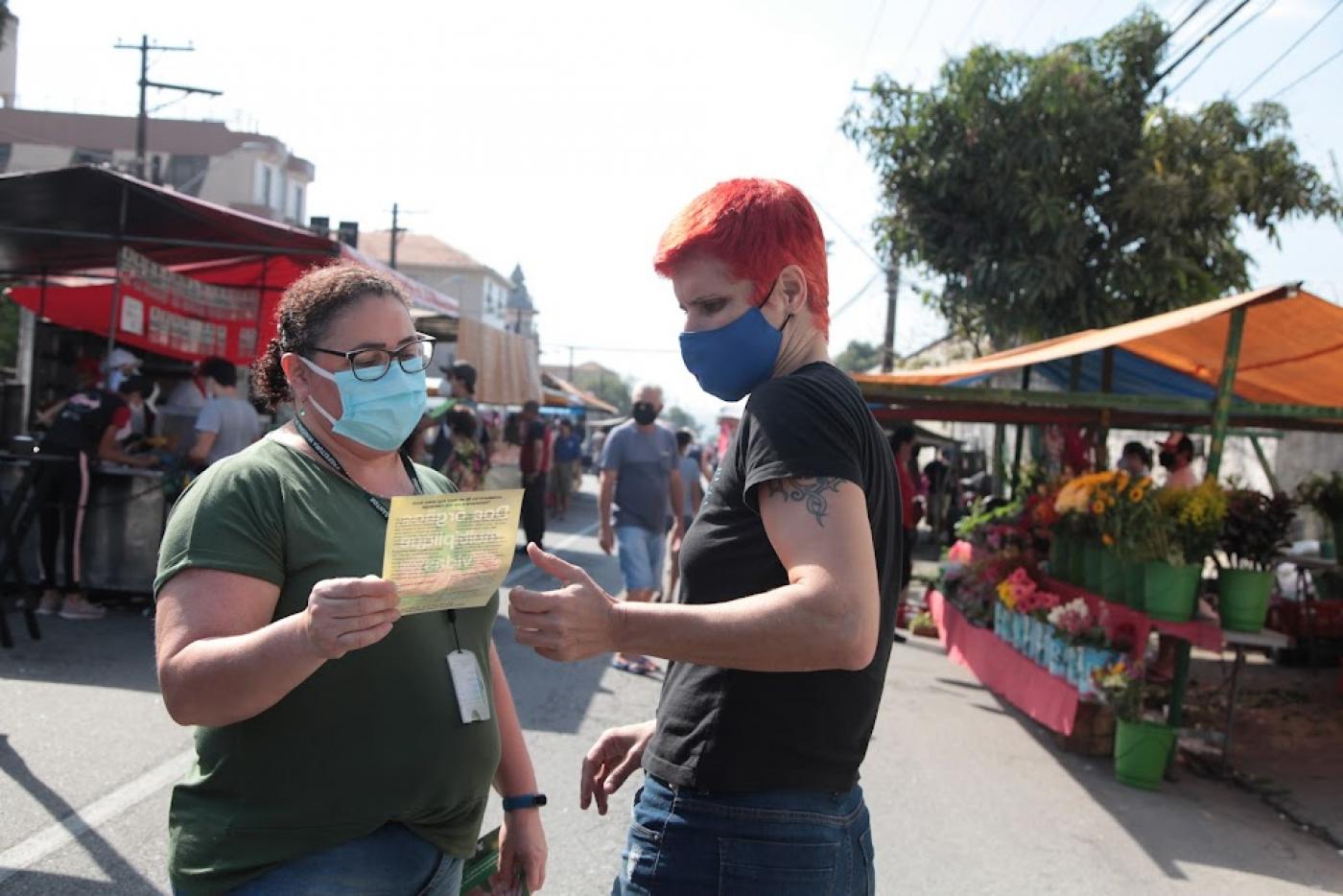 mulher entrega folheto a outra mulher no meio da feira. #paratodosverem
