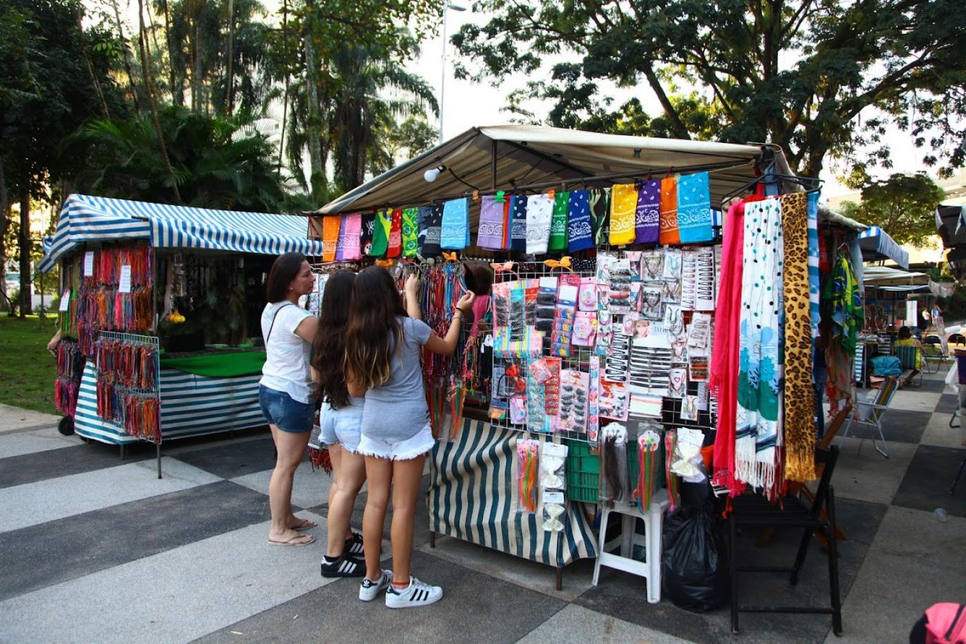 Três mulheres manuseiam artigos em barraca. #Paratodosverem